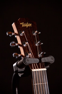 Close-up of a guitar's headstock with tuning pegs and strings, featuring a logo and mounted on a stand against a dark background.