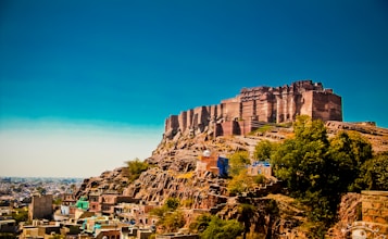 An ancient fort sits majestically atop a rocky hill, overlooking a city filled with colorful buildings. The sky is clear and blue, while lush green trees surround the base of the fort, adding to the vibrant landscape.