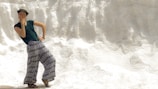 A person wearing a hat, dark green shirt, and patterned wide-legged pants strikes a playful pose against a textured, white background resembling a snowbank or salt mound. The person's expression and hand gesture convey a sense of fun and spontaneity.