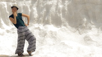 A person wearing a hat, dark green shirt, and patterned wide-legged pants strikes a playful pose against a textured, white background resembling a snowbank or salt mound. The person's expression and hand gesture convey a sense of fun and spontaneity.