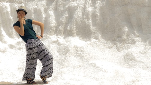 A person wearing a hat, dark green shirt, and patterned wide-legged pants strikes a playful pose against a textured, white background resembling a snowbank or salt mound. The person's expression and hand gesture convey a sense of fun and spontaneity.