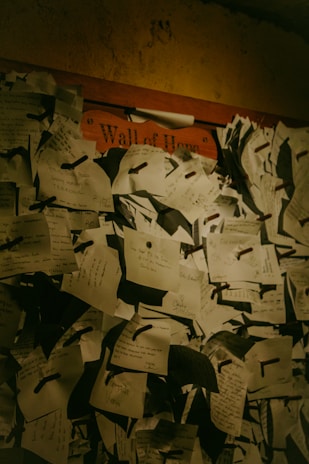 A dense cluster of handwritten notes pinned to a board, labeled 'Wall of Hope'. The notes are of various sizes and orientations, consisting of personal messages and aspirations. The lighting is dim, lending a warm, intimate feel to the scene.