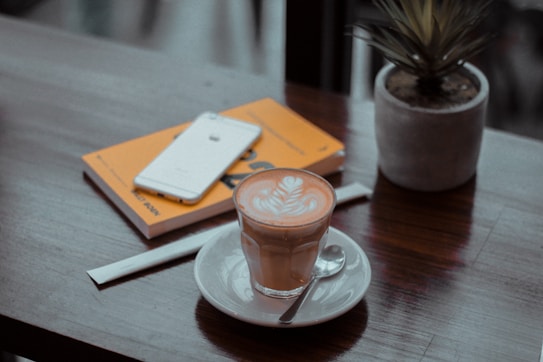A warm and cozy coffee shop setting with a latte on a white saucer placed on a wooden table. Next to the latte, there's a spoon and a smartphone resting on a closed book with a yellow cover. There is also a small potted plant adding a touch of greenery to the scene.