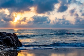 The image captures a serene beach scene at sunrise or sunset, with the sun partially obscured by clouds, casting a warm glow over the ocean. Waves gently lap against the rocky shoreline, creating a tranquil atmosphere.