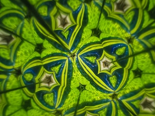 A vibrant, kaleidoscopic pattern featuring intricate geometric shapes. The design is dominated by rich hues of green and blue, forming symmetrical star-like and heart-like structures that create a mesmerizing, repetitive pattern.