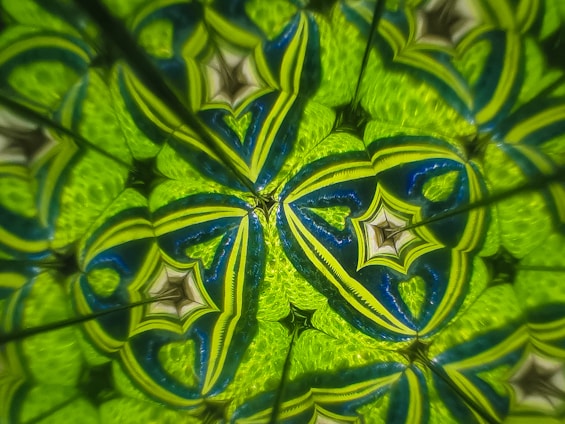 A vibrant, kaleidoscopic pattern featuring intricate geometric shapes. The design is dominated by rich hues of green and blue, forming symmetrical star-like and heart-like structures that create a mesmerizing, repetitive pattern.