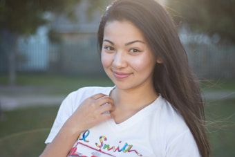 A young woman with long dark hair is smiling slightly, standing outdoors with greenery in the background. She is wearing a white T-shirt with colorful writing on it. Her expression is calm and pleasant, and the lighting suggests it is either morning or late afternoon.