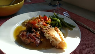 A well-lit dining table featuring a plate with a hearty meal. The dish includes a piece of roasted meat topped with cooked vegetables in a sauce. Green beans are placed beside the main dish, and a piece of crusty bread lies alongside. A fork is visible next to the plate, and a glass of red wine rests on the table in the background. The setting is casual with a focus on natural, warm lighting.