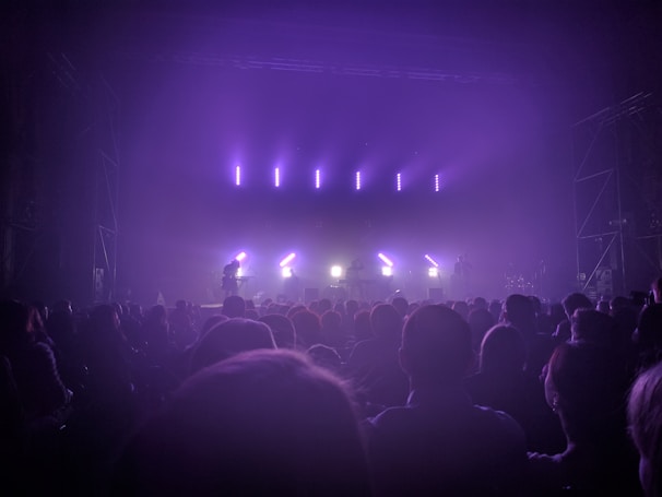A dimly lit concert venue features a stage with bright, purple and white lights shining through mist. The silhouettes of a band and musical equipment are visible in the background, while a large audience fills the foreground, creating a dynamic and lively atmosphere.