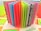 An assortment of colorful open pages in a notebook or scrapbook, with each page a different bright color or pattern. The background includes additional sheets or cards in various shades and patterns, including floral and leaf designs.