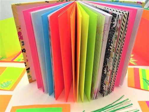 An assortment of colorful open pages in a notebook or scrapbook, with each page a different bright color or pattern. The background includes additional sheets or cards in various shades and patterns, including floral and leaf designs.