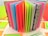 An assortment of colorful open pages in a notebook or scrapbook, with each page a different bright color or pattern. The background includes additional sheets or cards in various shades and patterns, including floral and leaf designs.