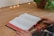 An open book with red and black text lies on a wooden table. Two hands are gently placed on either side of the book. A small string of warm, white lights runs along the edge of the table, creating a cozy ambiance. A potted plant with green leaves sits in the background.