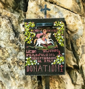 A donation box with an artistic depiction of a person on horseback slaying a dragon, surrounded by decorative floral patterns and inscriptions in multiple languages on its front. It is mounted against a rocky surface.