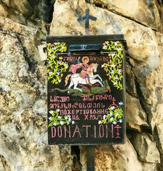 A donation box with an artistic depiction of a person on horseback slaying a dragon, surrounded by decorative floral patterns and inscriptions in multiple languages on its front. It is mounted against a rocky surface.