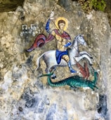 A richly detailed painting depicts a person dressed in medieval armor, riding a white horse. The figure holds a spear and appears to be engaged in combat with a green dragon-like creature. The background consists of textured stone, and there is ancient writing next to the figure.