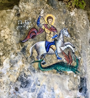 A richly detailed painting depicts a person dressed in medieval armor, riding a white horse. The figure holds a spear and appears to be engaged in combat with a green dragon-like creature. The background consists of textured stone, and there is ancient writing next to the figure.