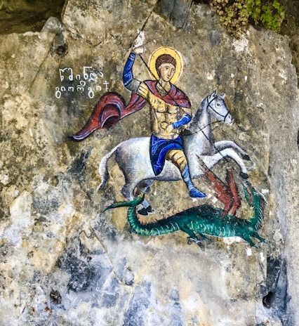 A richly detailed painting depicts a person dressed in medieval armor, riding a white horse. The figure holds a spear and appears to be engaged in combat with a green dragon-like creature. The background consists of textured stone, and there is ancient writing next to the figure.