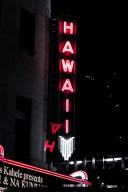 A vibrant neon blue and pink sign glowing against a Hawaiian beach sunset backdrop.