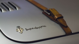 A close-up view of a metallic surface featuring the emblem and text 'Carrozzeria Touring Superleggera Milano.' Adjacent to the emblem is a leather strap with a buckle, adding a vintage or luxury feel to the image.