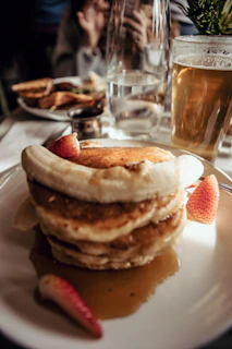 A stack of pancakes is topped with banana slices and strawberries, served on a white plate with syrup. A glass of beer is placed to the right, and there are blurred backgrounds of other dishes and a person.