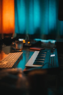 A dimly lit room featuring a keyboard synthesizer and a computer keyboard on a desk. The background includes softly glowing orange and teal curtains, creating a cozy and creative atmosphere.