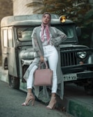 A woman stands confidently in front of an old, rugged vehicle. She is dressed in a stylish gray duster, white pants, and high heels. A soft pink scarf and matching handbag complement her outfit. Her makeup is bold with pink eyeshadow. The scene exudes a blend of urban and vintage aesthetics, with the woman striking a fashionable pose.