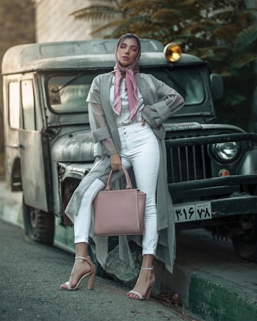 A woman stands confidently in front of an old, rugged vehicle. She is dressed in a stylish gray duster, white pants, and high heels. A soft pink scarf and matching handbag complement her outfit. Her makeup is bold with pink eyeshadow. The scene exudes a blend of urban and vintage aesthetics, with the woman striking a fashionable pose.