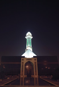 A mosque with a tall minaret illuminated at night. The structure features intricate architectural details with a prominent green light shining from the minaret. The front entrance is ornately decorated and reflects in a pool of water.