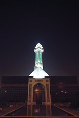 A mosque with a tall minaret illuminated at night. The structure features intricate architectural details with a prominent green light shining from the minaret. The front entrance is ornately decorated and reflects in a pool of water.