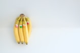 Four bananas are grouped together with small decorative eyes and colorful balls attached, making them resemble playful characters on a plain, light-colored background.