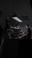 A dimly lit cafe space with a focus on a barista working at an espresso machine. The setting is modern and minimalist, with a prominent hanging light illuminating the work area. There are multiple coffee-making tools and containers placed on the counter.