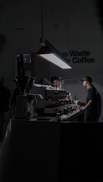 A dimly lit cafe space with a focus on a barista working at an espresso machine. The setting is modern and minimalist, with a prominent hanging light illuminating the work area. There are multiple coffee-making tools and containers placed on the counter.