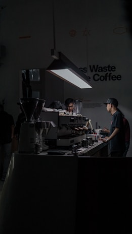 A dimly lit cafe space with a focus on a barista working at an espresso machine. The setting is modern and minimalist, with a prominent hanging light illuminating the work area. There are multiple coffee-making tools and containers placed on the counter.