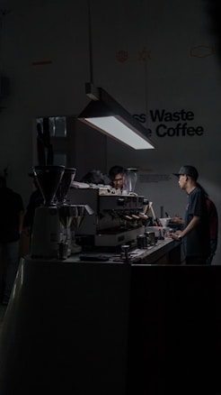 A dimly lit cafe space with a focus on a barista working at an espresso machine. The setting is modern and minimalist, with a prominent hanging light illuminating the work area. There are multiple coffee-making tools and containers placed on the counter.
