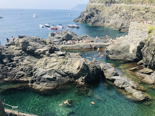 Crystal-clear turquoise waters surround large rocky formations along a rugged coastline. Numerous people are swimming, sunbathing, and enjoying the warm weather. Small boats are anchored in the distance on the calm sea.