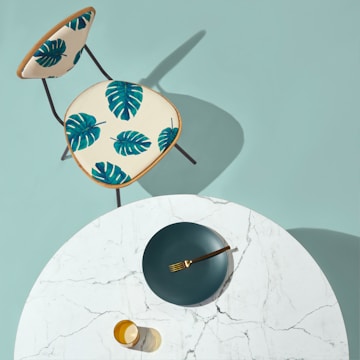 A modern, minimalist setup with a round white marble table and a gold-colored fork resting on a dark green plate. A transparent amber glass sits nearby. Next to the table is a chair with a cream-colored seat featuring vibrant green monstera leaf patterns.