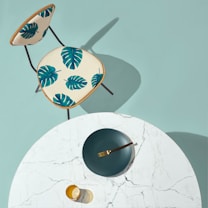 A modern, minimalist setup with a round white marble table and a gold-colored fork resting on a dark green plate. A transparent amber glass sits nearby. Next to the table is a chair with a cream-colored seat featuring vibrant green monstera leaf patterns.