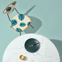 A modern, minimalist setup with a round white marble table and a gold-colored fork resting on a dark green plate. A transparent amber glass sits nearby. Next to the table is a chair with a cream-colored seat featuring vibrant green monstera leaf patterns.