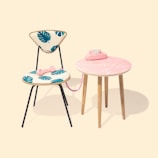 A mid-century modern chair with vibrant blue leaf patterns on its cushioned seat and backrest, paired with a small, round pink table featuring a marble-like finish. A vintage pink rotary telephone is placed on the table, with the receiver resting on the chair's seat. The setting is minimalistic with a soft, pastel background.