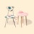A mid-century modern chair with vibrant blue leaf patterns on its cushioned seat and backrest, paired with a small, round pink table featuring a marble-like finish. A vintage pink rotary telephone is placed on the table, with the receiver resting on the chair's seat. The setting is minimalistic with a soft, pastel background.