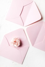 Soft blush pink envelope with a delicate wedding ring beside it on a wooden table.