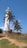A tall white lighthouse stands prominently against a clear blue sky, surrounded by swaying palm trees. A path leads to the lighthouse, bordered by grass and a small stone wall. The scene is bright and peaceful.