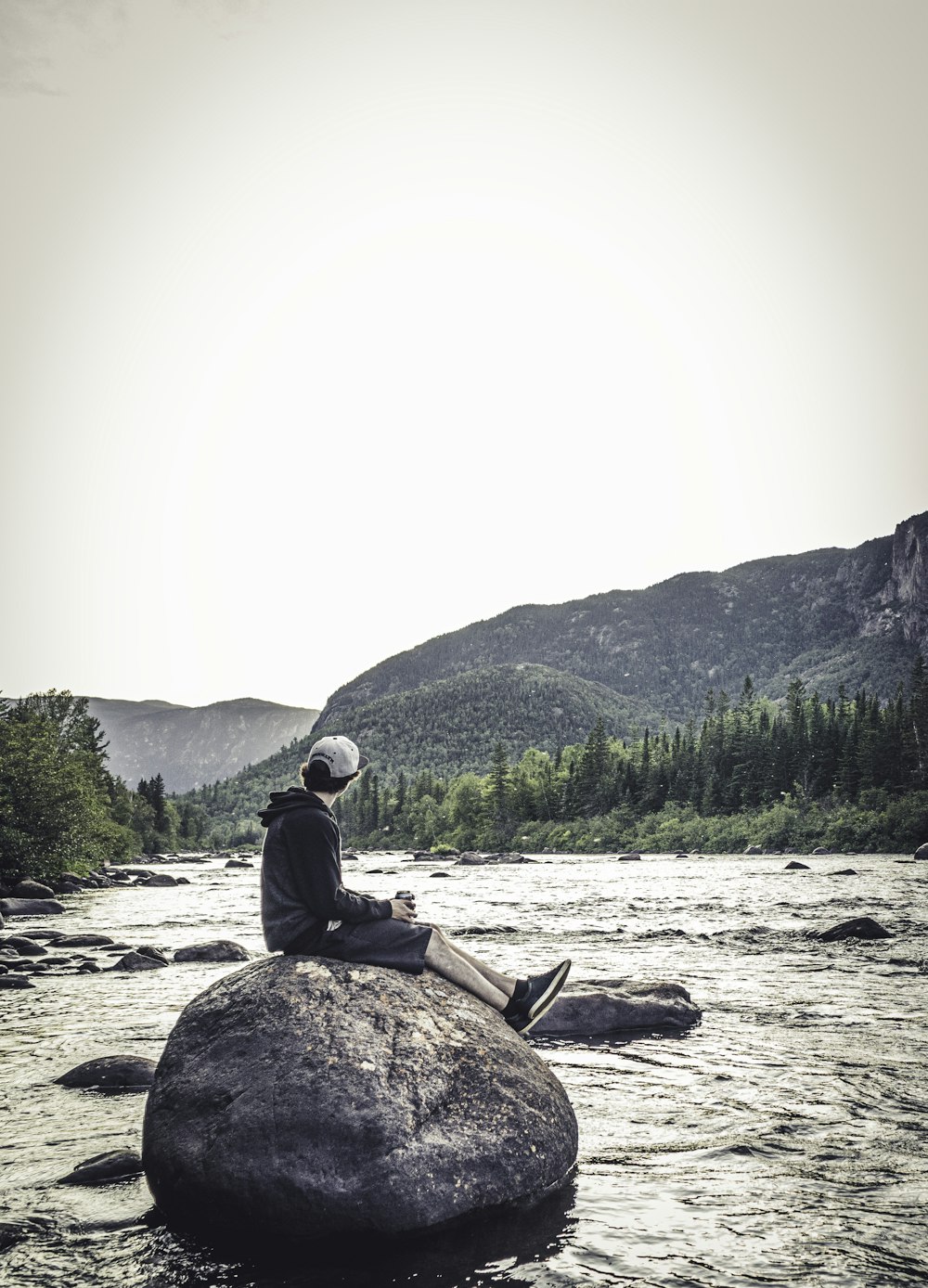 man sitting on rock viewing river and mountain during daytime
