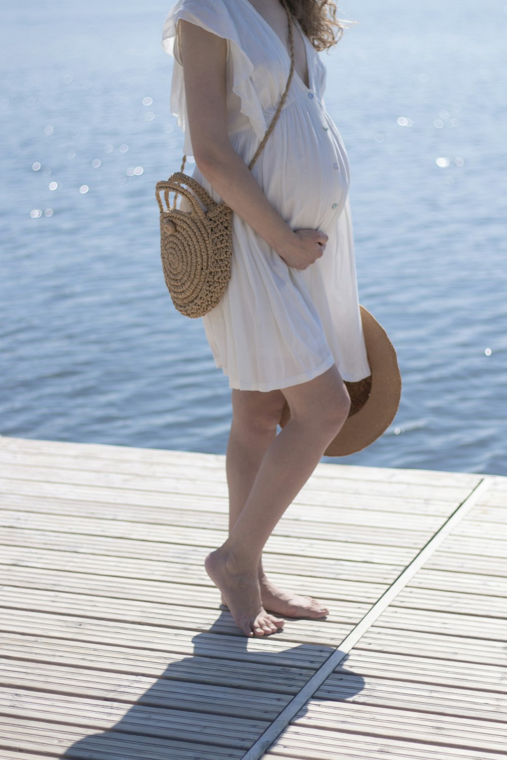 pregnant woman touching her belly and holding her hat standing on wooden dock during day