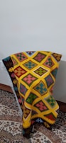 A colorful woven textile with intricate geometric patterns in shades of yellow, orange, red, blue, green, and pink is draped over a chair positioned against a plain wall. The textile features distinct diamond shapes and other symmetric motifs, creating a vibrant and eye-catching appearance. It rests on a traditional, ornately patterned rug with complex floral designs.