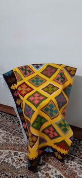 A colorful woven textile with intricate geometric patterns in shades of yellow, orange, red, blue, green, and pink is draped over a chair positioned against a plain wall. The textile features distinct diamond shapes and other symmetric motifs, creating a vibrant and eye-catching appearance. It rests on a traditional, ornately patterned rug with complex floral designs.