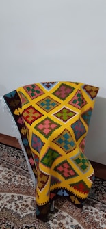 A colorful woven textile with intricate geometric patterns in shades of yellow, orange, red, blue, green, and pink is draped over a chair positioned against a plain wall. The textile features distinct diamond shapes and other symmetric motifs, creating a vibrant and eye-catching appearance. It rests on a traditional, ornately patterned rug with complex floral designs.