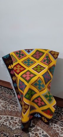A colorful woven textile with intricate geometric patterns in shades of yellow, orange, red, blue, green, and pink is draped over a chair positioned against a plain wall. The textile features distinct diamond shapes and other symmetric motifs, creating a vibrant and eye-catching appearance. It rests on a traditional, ornately patterned rug with complex floral designs.