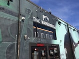 A brick building painted with a large mural featuring musical instruments and the silhouette of a person playing a guitar. The building has a sign reading 'The Warehouse' with the Perrier logo above it. There are industrial lights mounted on the wall and a glass entrance indicating entry and exit doors.